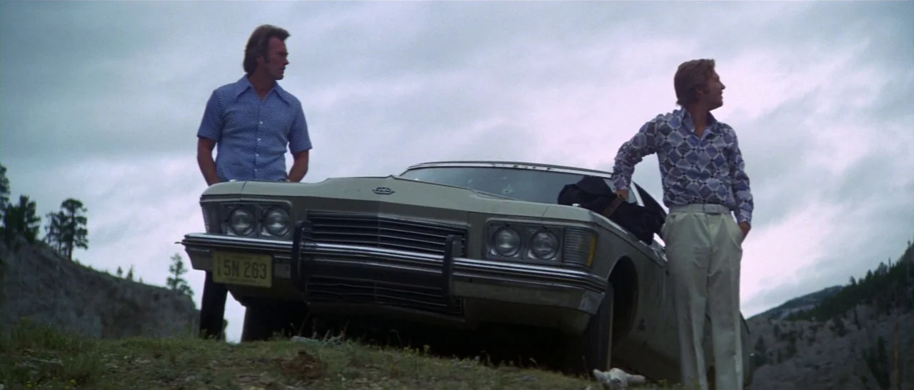 Thunderbolt & Lightfoot (1974) movie still: wide — A silver vintage sedan sits on a grassy, rocky hillside under an overcast sky. Two men stand…