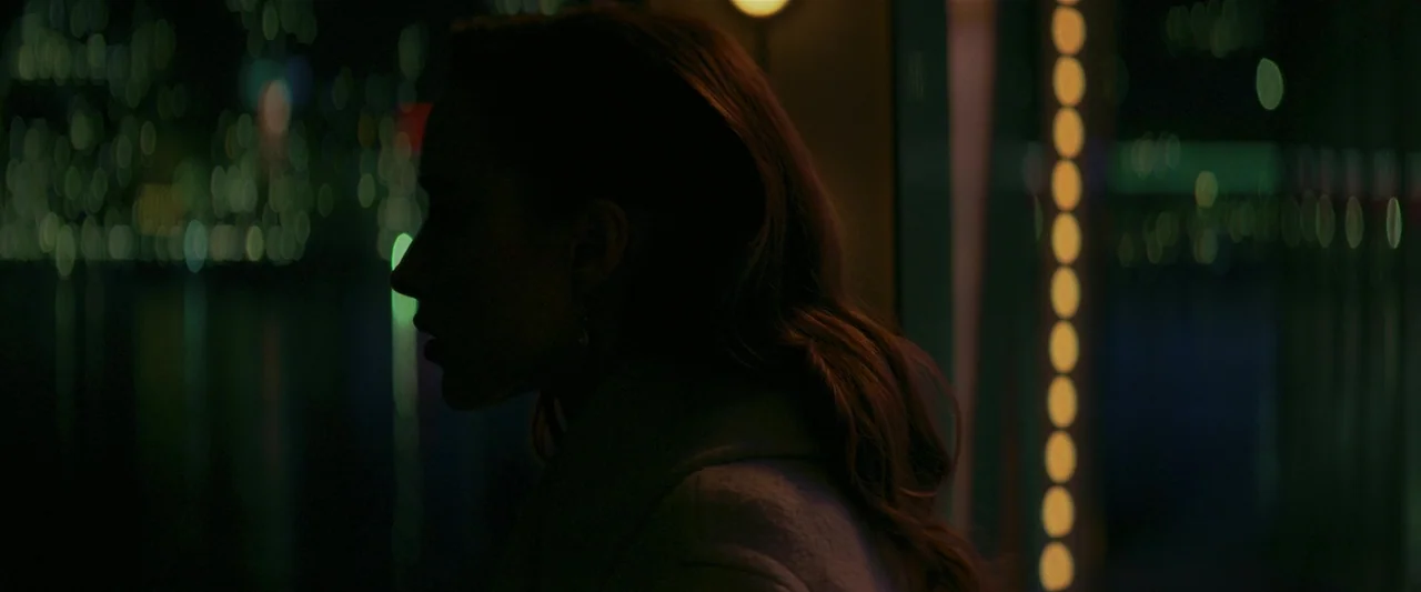 Fair Play (2023) movie still: closeup — A woman stands on an outdoor balcony at night with city lights behind her. She faces sideways while looking…
