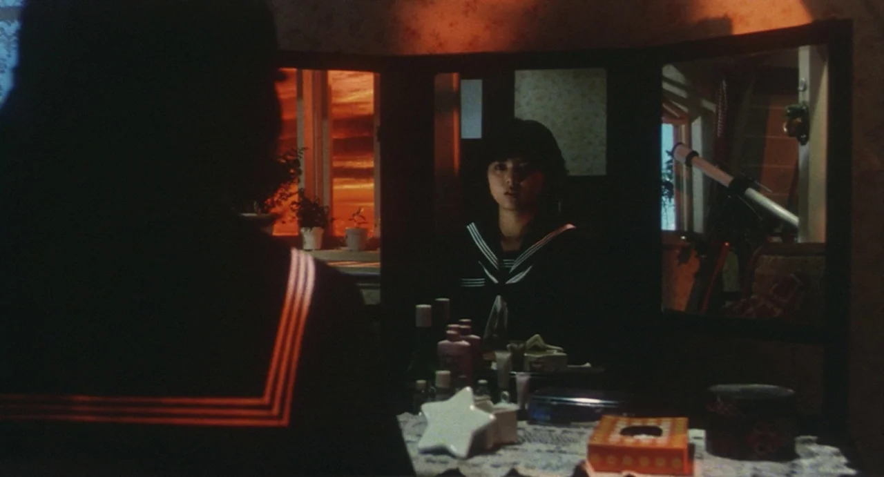 School in the Crosshairs (1981) movie still: medium — A dimly lit room features a wooden vanity with a three-paneled mirror. A young woman in a…