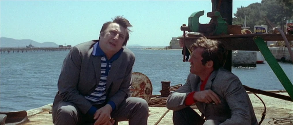 Pierrot Le Fou (Crazy Pete) (1965) movie still: medium — A wooden pier extends over bright blue water under a clear sunny sky. Two men in suits…