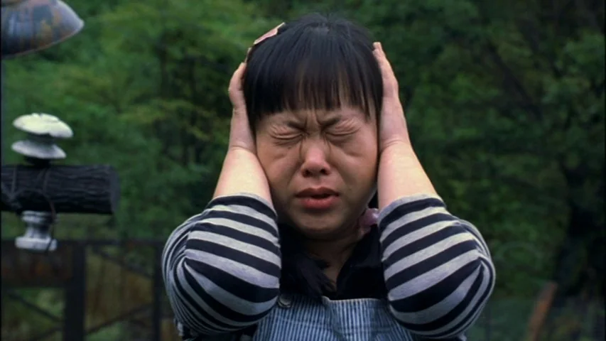 Save The Green Planet! (2003) movie still: closeup — The outdoor setting is a wooded area with trees visible behind the subject. A woman holds her…