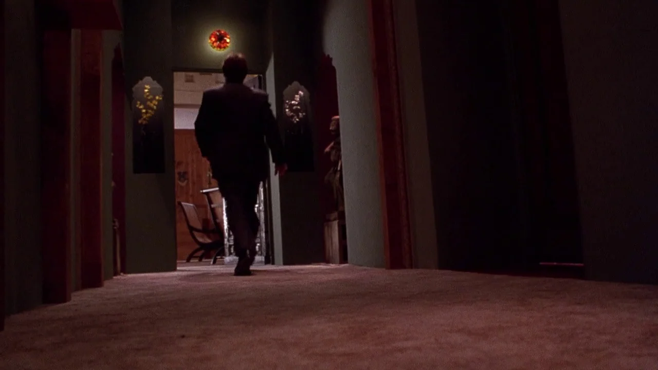 Secretary movie still: fullbody — A dark, carpeted hotel hallway features deep green walls and a single lit sconce. A man in a dark suit walks away…