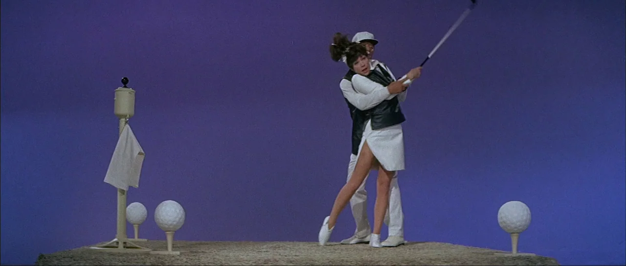 What A Way To Go! (1964) movie still: fullbody — A surreal studio stage with a solid violet background and large oversized golf props. An older man…