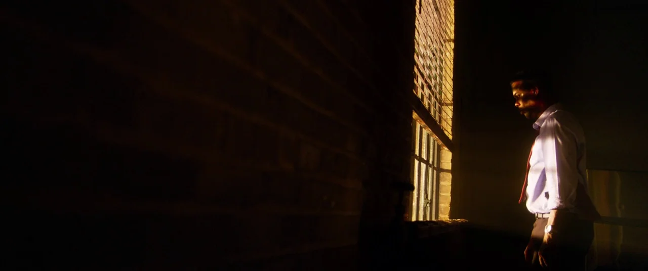 Spiral (2021) movie still: medium — An industrial space with a dark exposed brick wall and a large window. A man in a dress shirt stands by the…