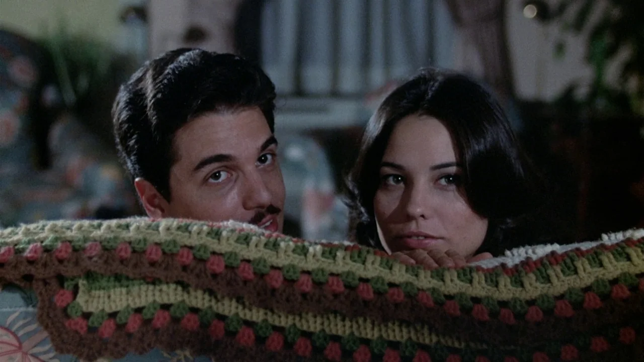 The Sentinel (1977) movie still: medium — A man and a woman rest behind a crocheted afghan blanket in a dark interior. They gaze directly toward the…