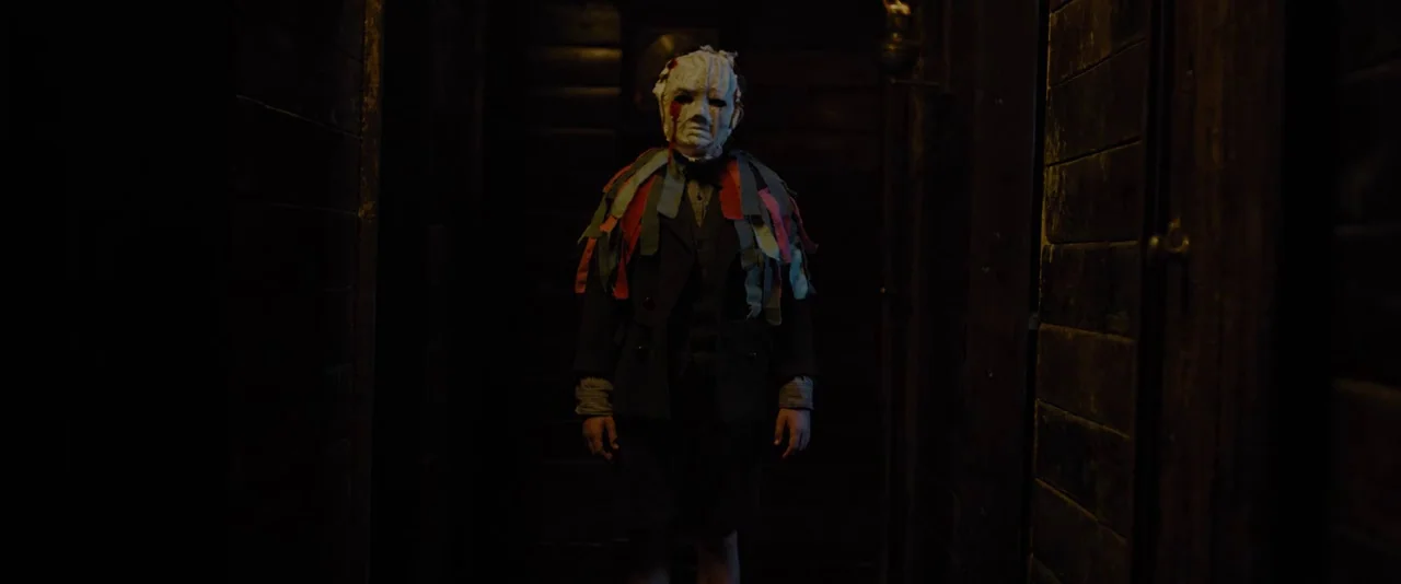 Apostle (2018) movie still: fullbody — A dark, cramped wooden hallway features horizontal planks and low-key illumination. A child wearing a white…