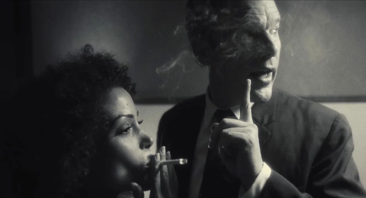 Born To Be Blue (2015) movie still: medium — A dimly lit room with dark walls and heavy shadows. A man signals for silence while a woman holds a…