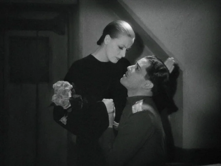 Mata Hari (1931) movie still: medium — A dimly lit staircase area provides the setting for the two figures. A man kneels before a woman holding a…