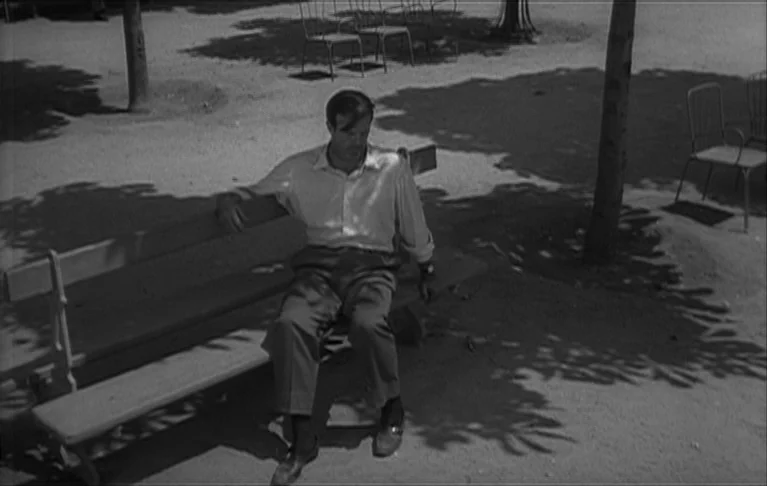 Le Signe du Lion (1962) movie still: fullbody — A gravel-covered park area features scattered metal chairs and bare tree trunks. A man wearing a…