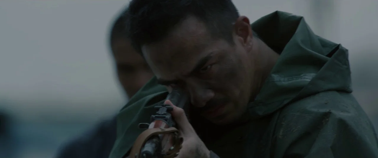 The Night Comes for Us (2018) movie still: closeup — A field under a dull, overcast sky provides a muted outdoor setting. A man in a green poncho…