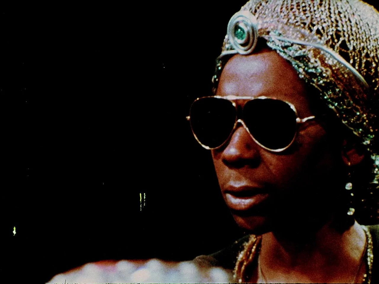 Space is the Place (1974) movie still: closeup — A dark environment with a woman wearing an elaborate gold headpiece and sunglasses. The woman…