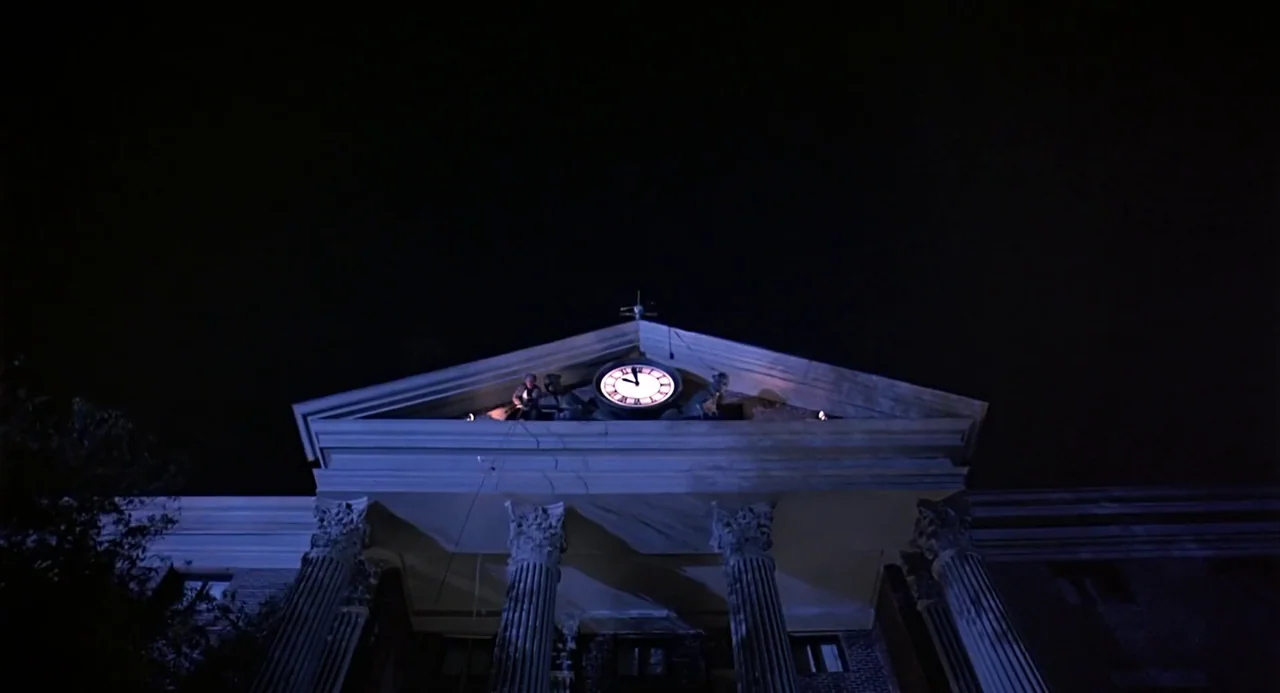 Back to the Future (1985) movie still: wide — A classical stone pediment with columns supports a large glowing clock at night. The architecture is…