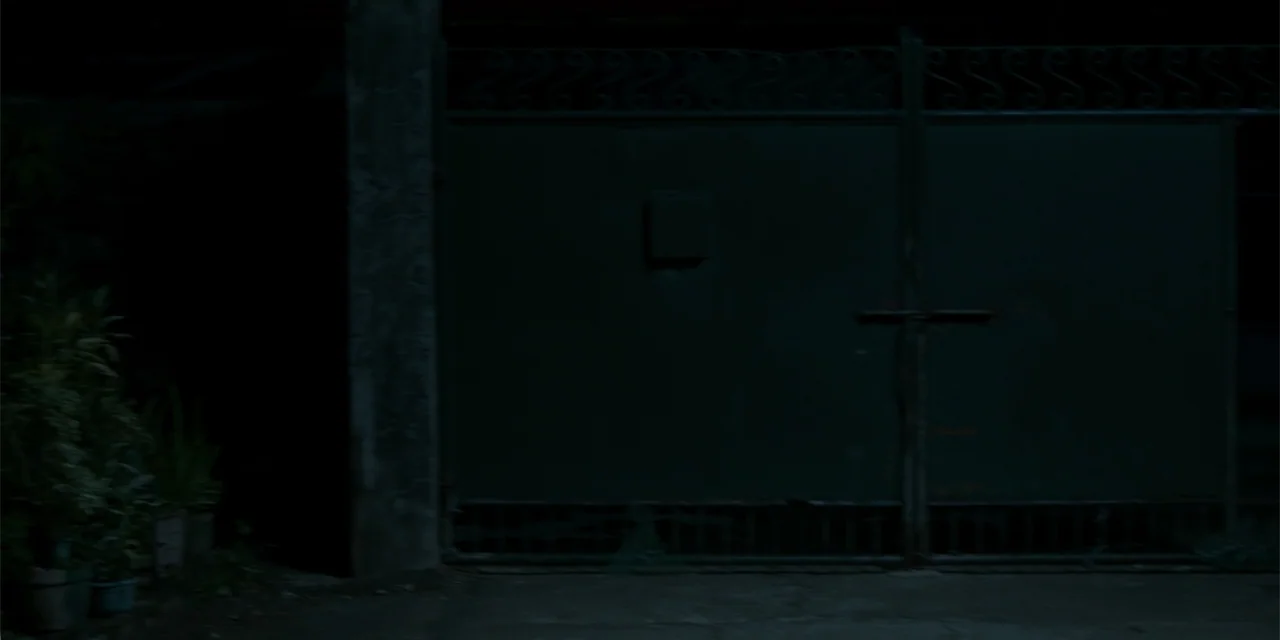 Nokturno (2024) movie still: establishing — A dark night scene features a closed iron gate flanked by potted plants. The structure stands in deep…