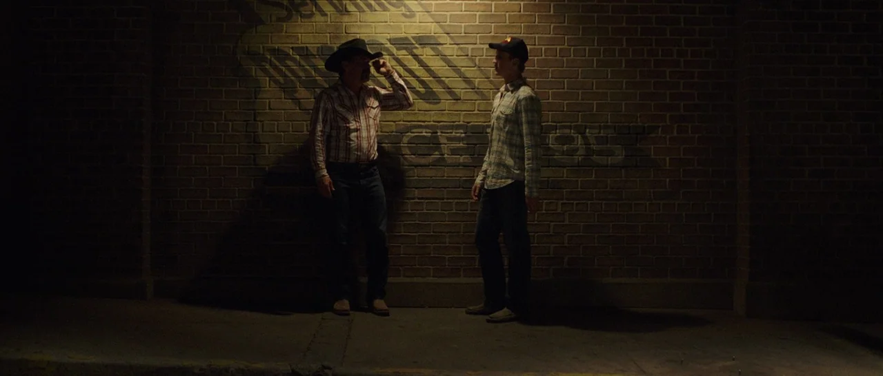 Only The Brave (2017) movie still: fullbody — A dark, exterior location features a large brown brick wall with a painted logo. Two men stand in a…