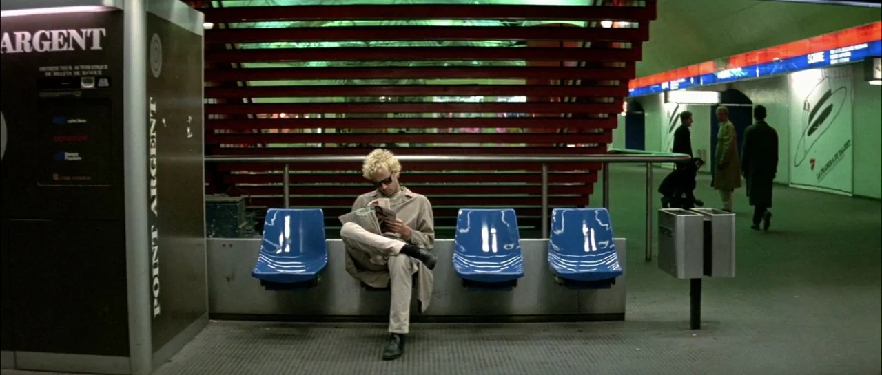 Subway (1985) movie still: wide — A modern transit terminal features bright blue seats set against red slats. A solitary man sits reading a…