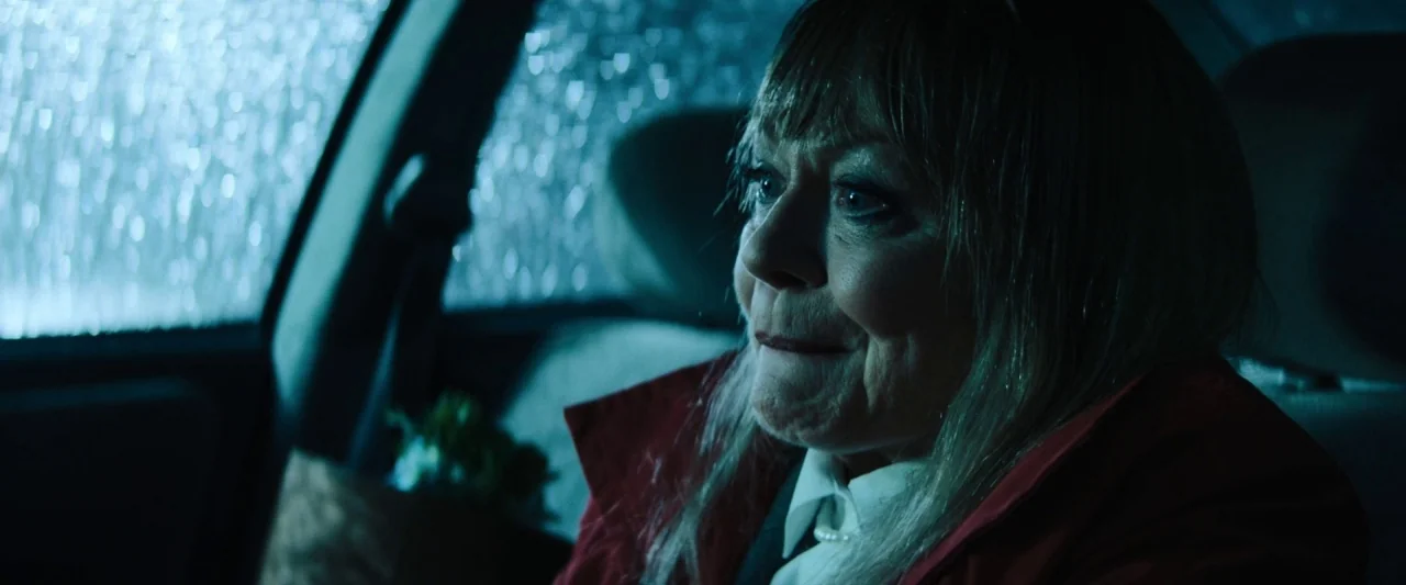 The Grudge (2020) movie still: closeup — A woman sits inside a car at night while rain streaks the side window. She looks toward the glass with a…