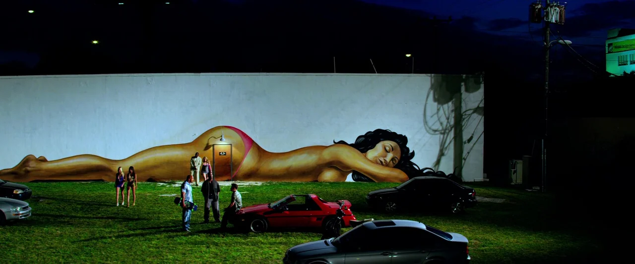 Pain & Gain (2013) movie still: wide — A large mural of a reclining woman is painted on a white wall. Several people stand on a patch of grass in…