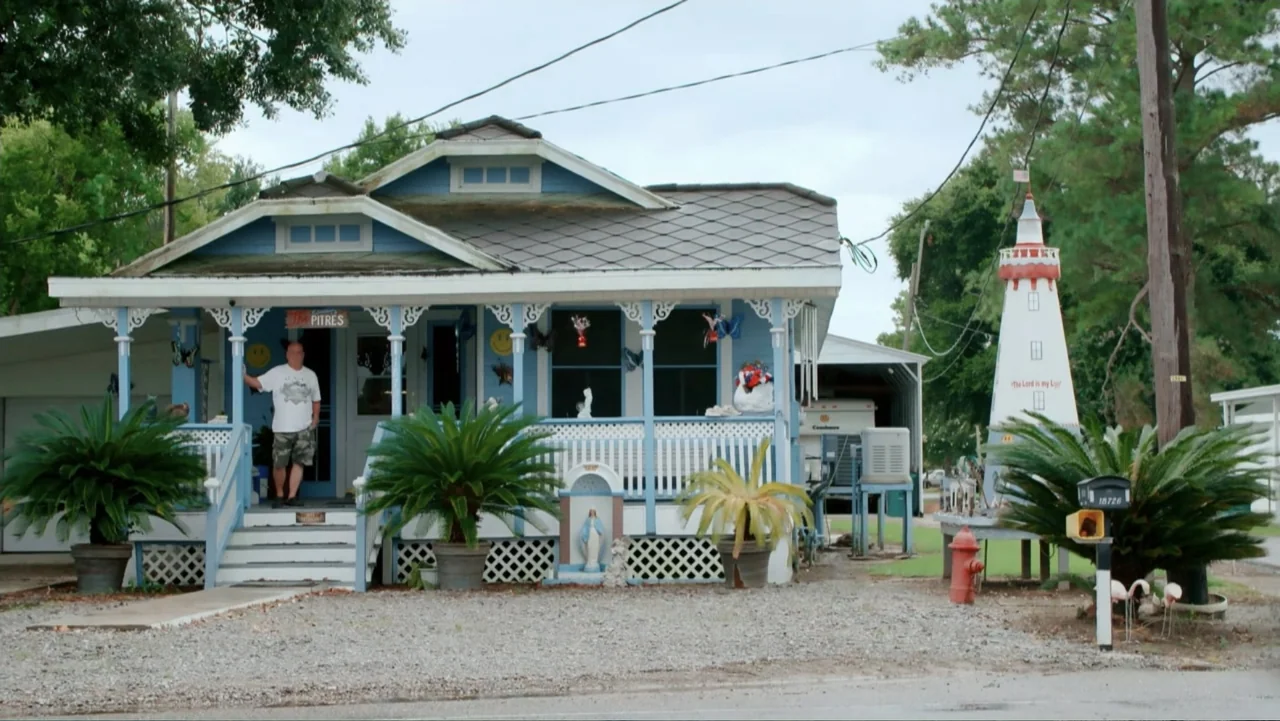 Mommy Dead and Dearest (2017) movie still: wide — A blue suburban house with a white porch railing features a lighthouse yard ornament nearby. A man…