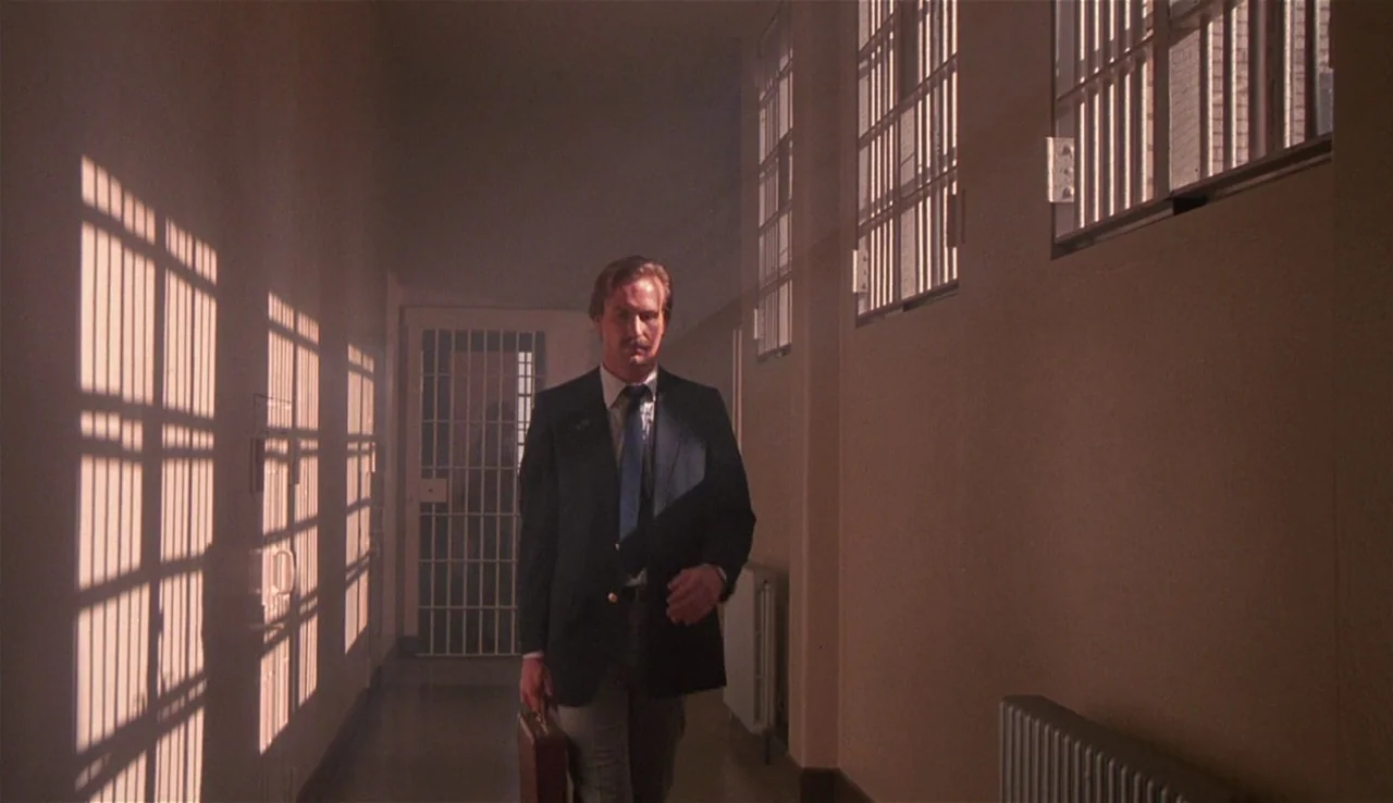 Body Heat (1981) movie still: fullbody — A long interior hallway features high walls, barred windows, and metal gated doors. A man in a business…