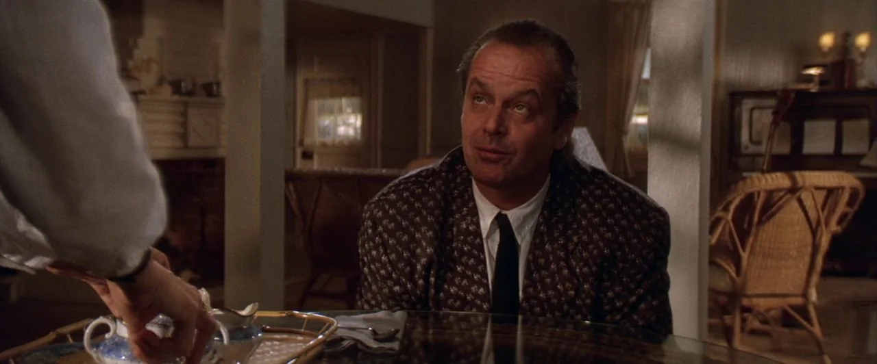 The Witches of Eastwick (1987) movie still: medium — A residential interior features wicker chairs and a glass-topped table. A man in a blazer sits…