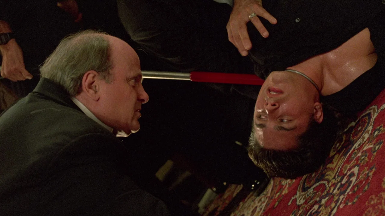 Society (1989) movie still: medium — A dimly lit interior with a dark red patterned rug covering the floor. An older man observes a man lying on the…