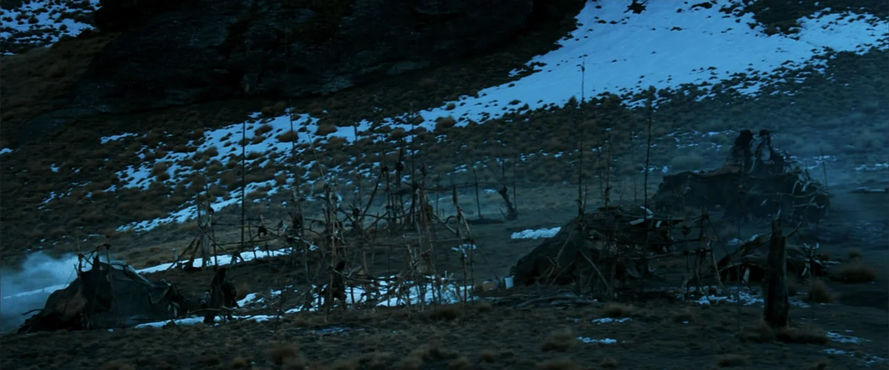 10,000 BC (2008) movie still: wide — A remote wilderness encampment features crude wooden frames and low shelters on a slope. Patches of snow cling…