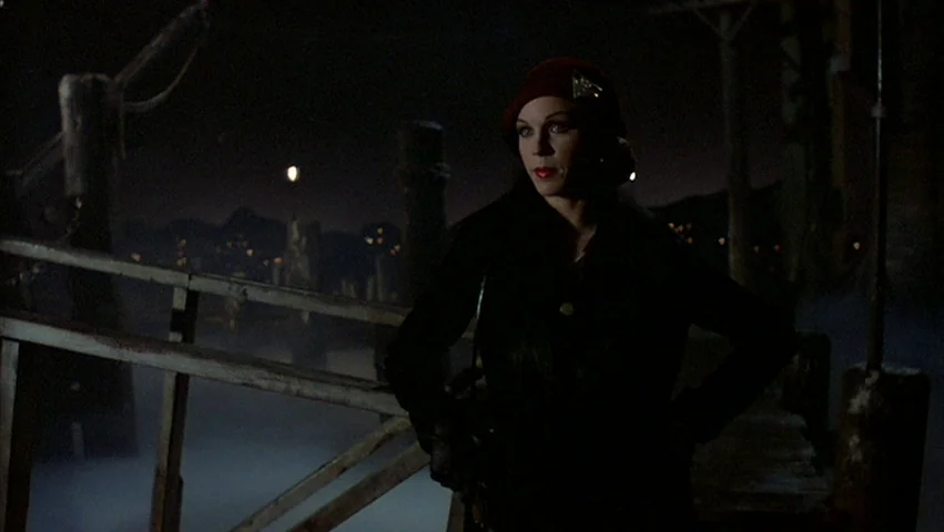 Hammett (1982) movie still: medium — A wooden pier at night with fog rising from the dark water below. A woman in a dark coat and red beret stands…
