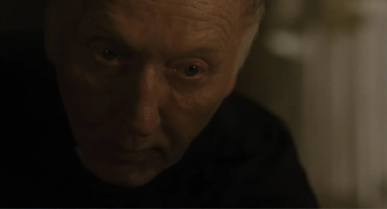 Gates of Darkness (2019) movie still: closeup — A dark indoor space with soft lighting focused on a man's face. An elderly man with grey hair looks…