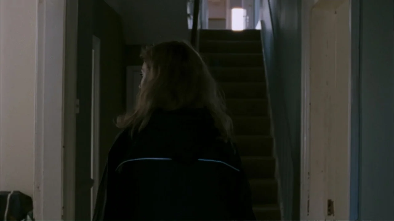 28 Weeks Later (2007) movie still: medium — A narrow residential hallway leads to a staircase illuminated by a single distant light. A woman walks…