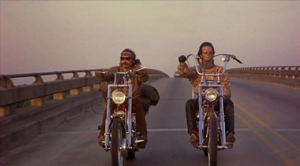 Easy Rider (1969) movie still: fullbody — A long concrete bridge stretches toward the horizon under a hazy sky. Two men ride custom motorcycles…