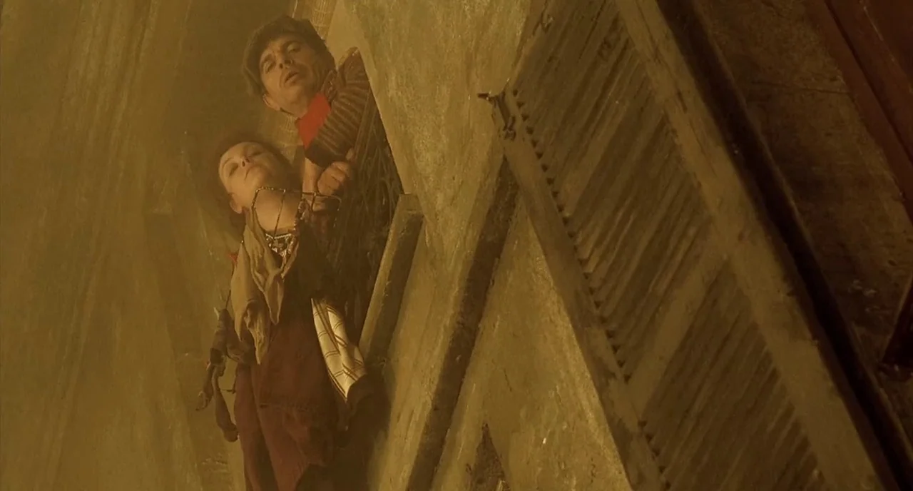 Delicatessen (1991) movie still: medium — A stone building exterior features a wooden window shutter and an open window frame. A man and woman gaze…
