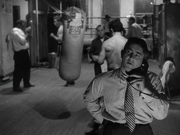Killer’s Kiss (1955) movie still: medium — A dark, utilitarian training facility features a large hanging heavy bag and ring ropes. A man in a dress…