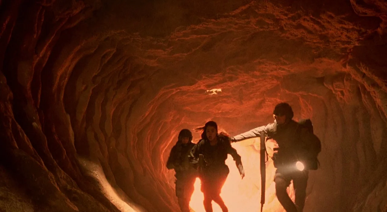 Starship Troopers (1997) movie still: wide — A narrow, textured rock tunnel glows with an intense orange light from below. Three people in tactical…