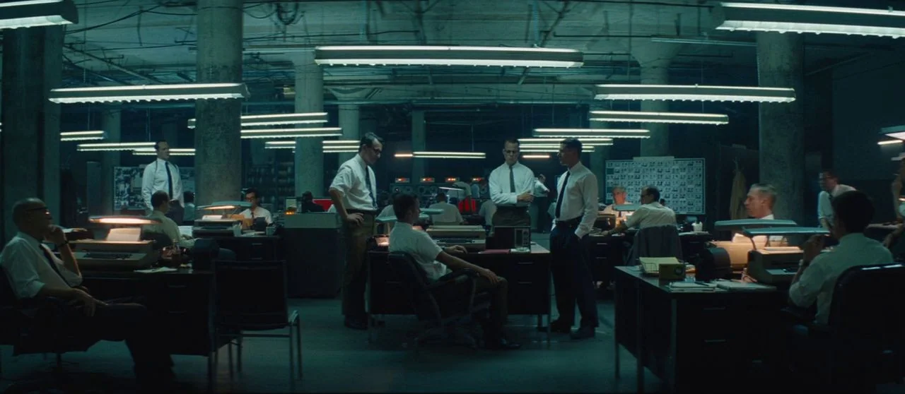 Seberg (2019) movie still: wide — A spacious, dimly lit mid-century workspace is filled with rows of desks and staff. Men in professional attire are…