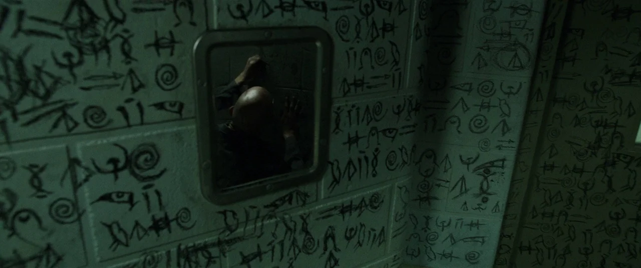 Shazam! (2019) movie still: medium — A concrete wall in a room is covered in extensive black hand-drawn cryptic symbols. A man stands facing the…