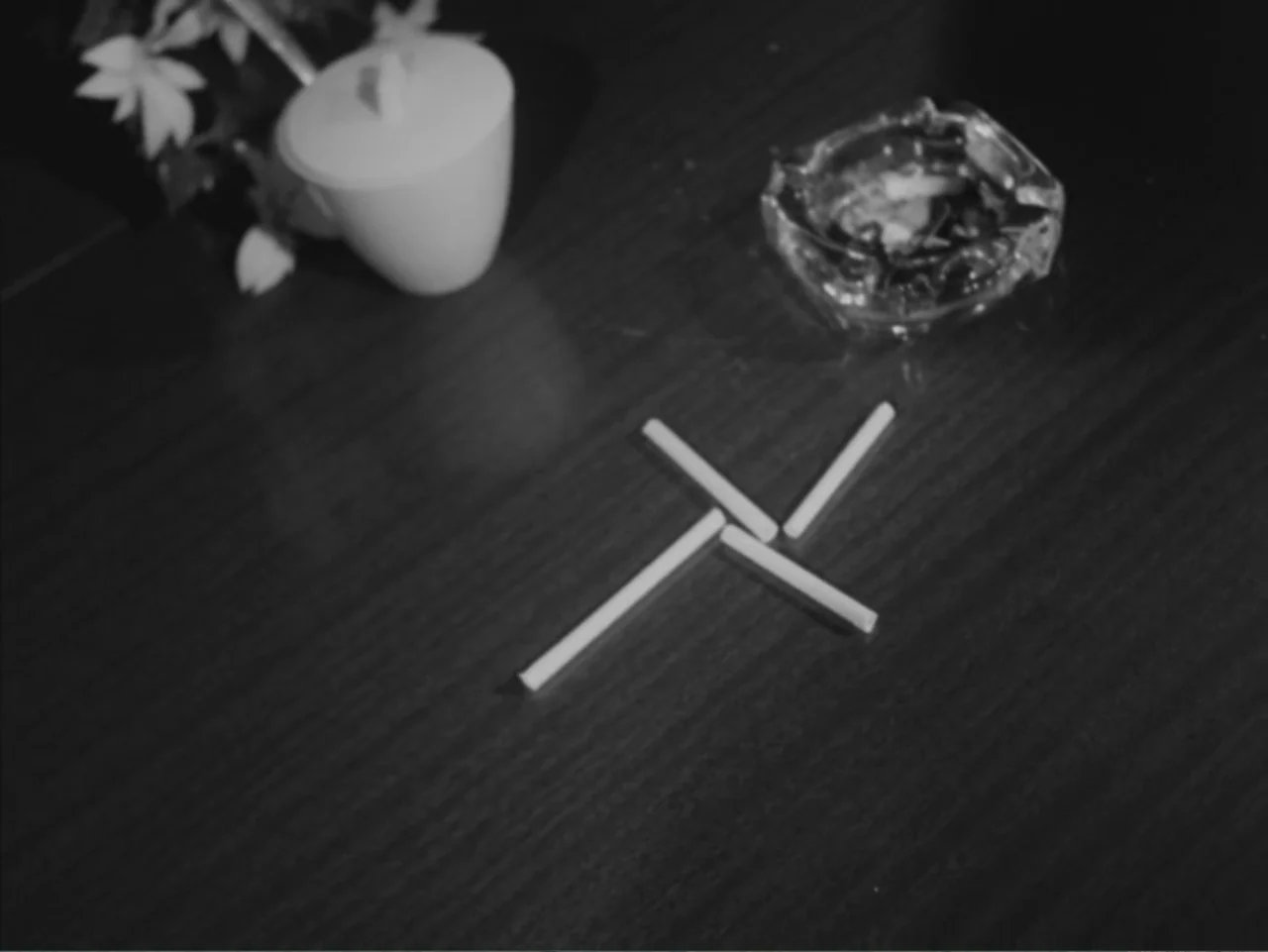 Chained Girls (1965) movie still: closeup — A dark wooden surface holds an arranged pattern of cigarettes next to a glass ashtray. The cigarettes…