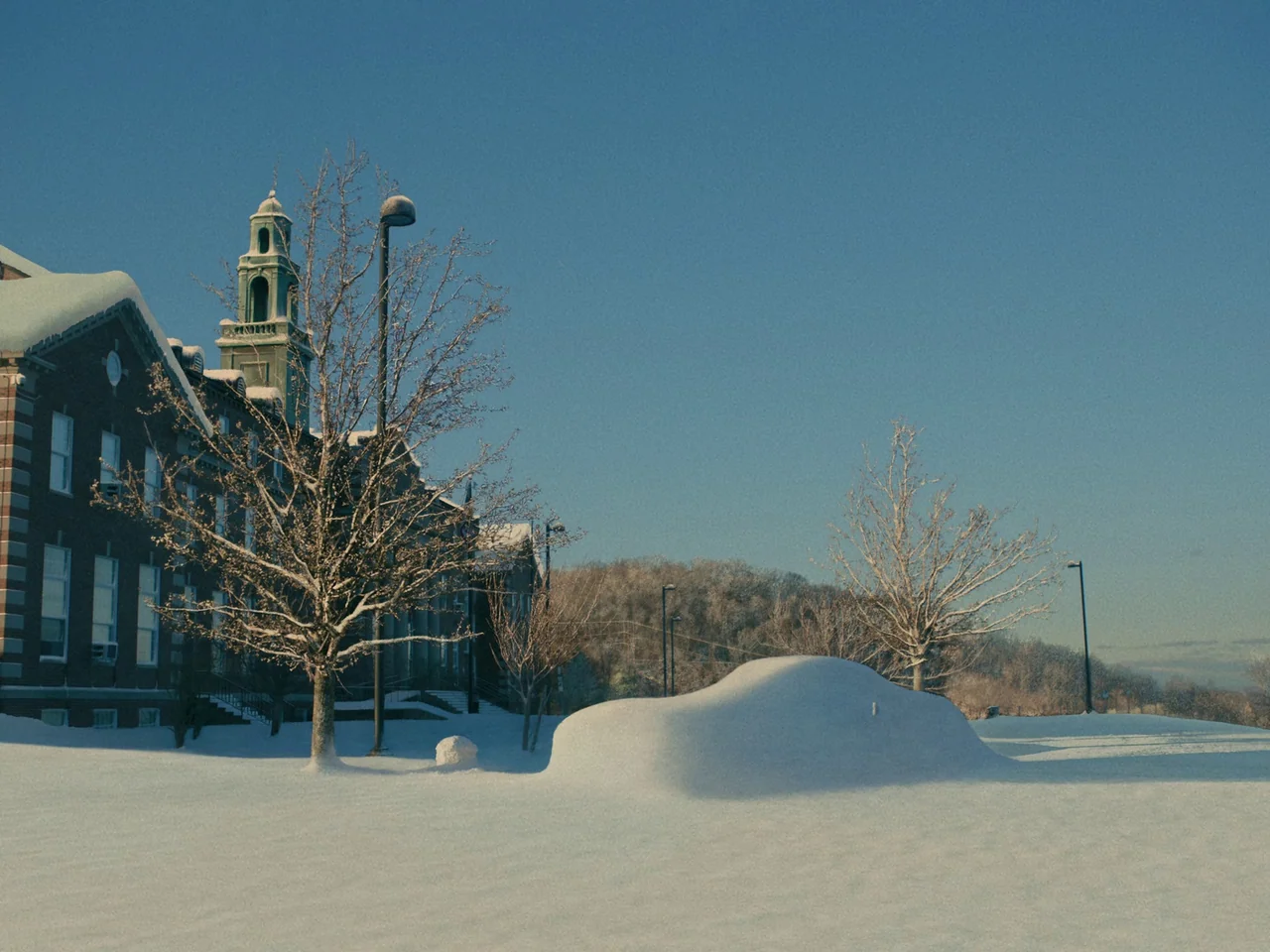 I’m Thinking of Ending Things (2020) movie still: wide — A brick building with a bell tower stands beside a field covered in thick snow. A single…