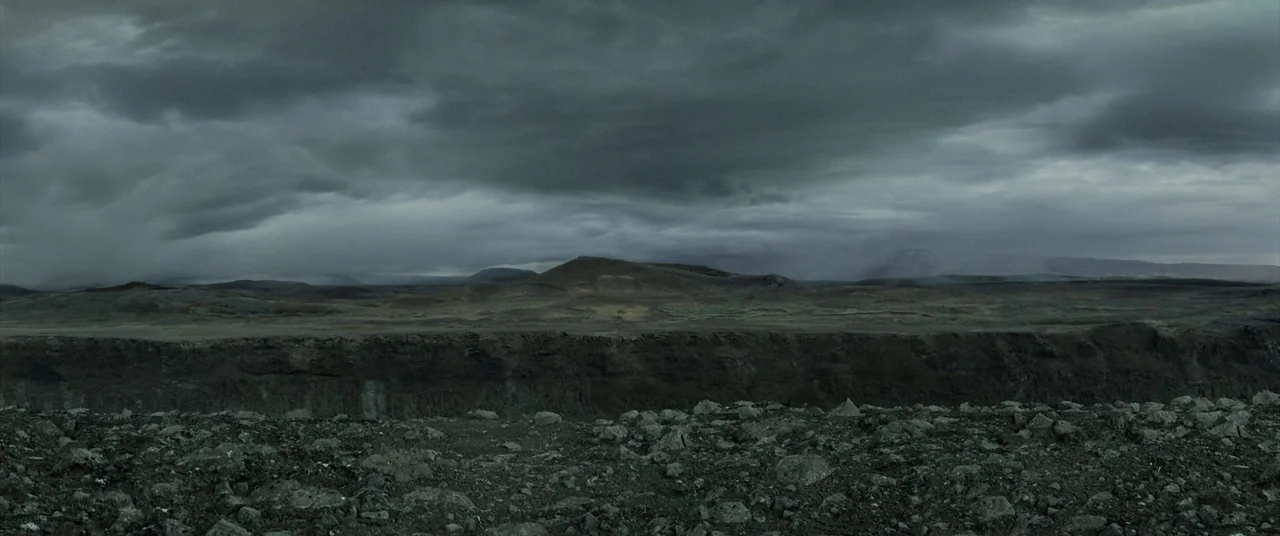 Oblivion (2013) movie still: establishing — A desolate and expansive volcanic landscape stretches under a thick, low-hanging overcast sky. Dark…