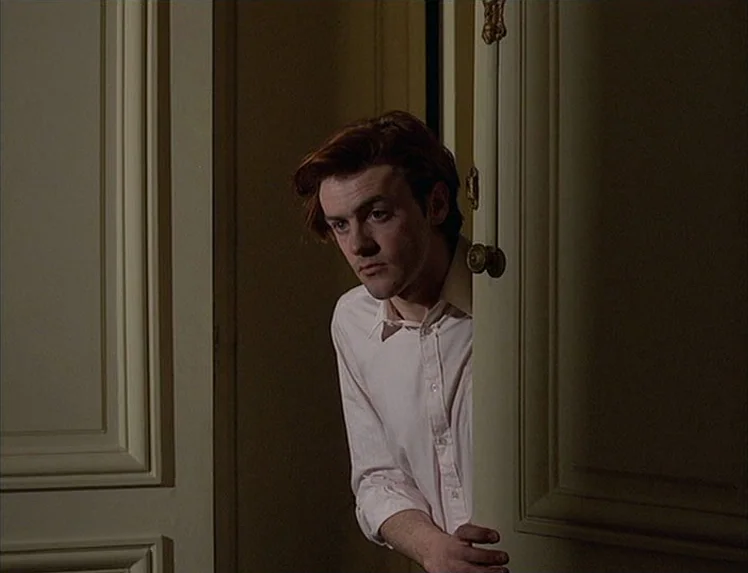 First Name: Carmen (1983) movie still: medium — A person stands in a dimly lit doorway with classic molding and white panels. A man in a white dress…