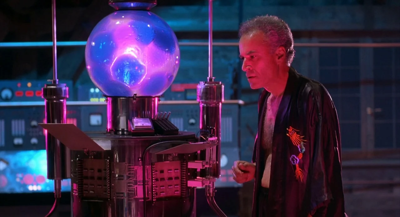 From Beyond (1986) movie still: medium — A dimly lit room contains a metallic machine topped with a glowing plasma globe. An older man in a silk…