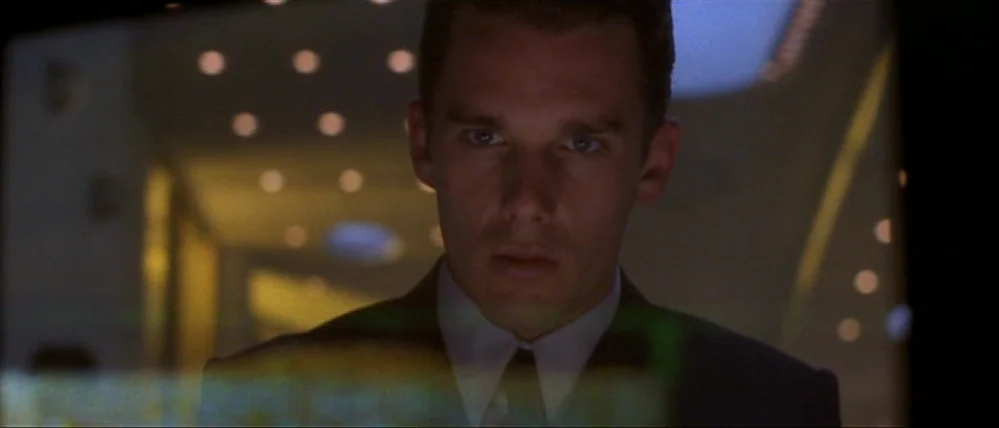 Gattaca (1997) movie still: closeup — A man in a dark suit sits in a darkened office at night. He is staring directly at a bright screen reflecting…