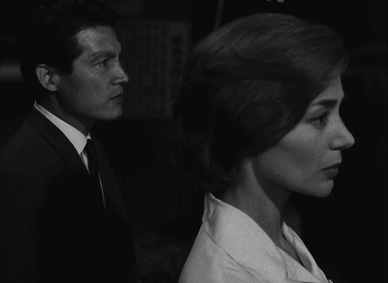 Hiroshima Mon Amour (1959) movie still: medium — A dimly lit indoor space with a completely black background. A man and a woman are positioned in…