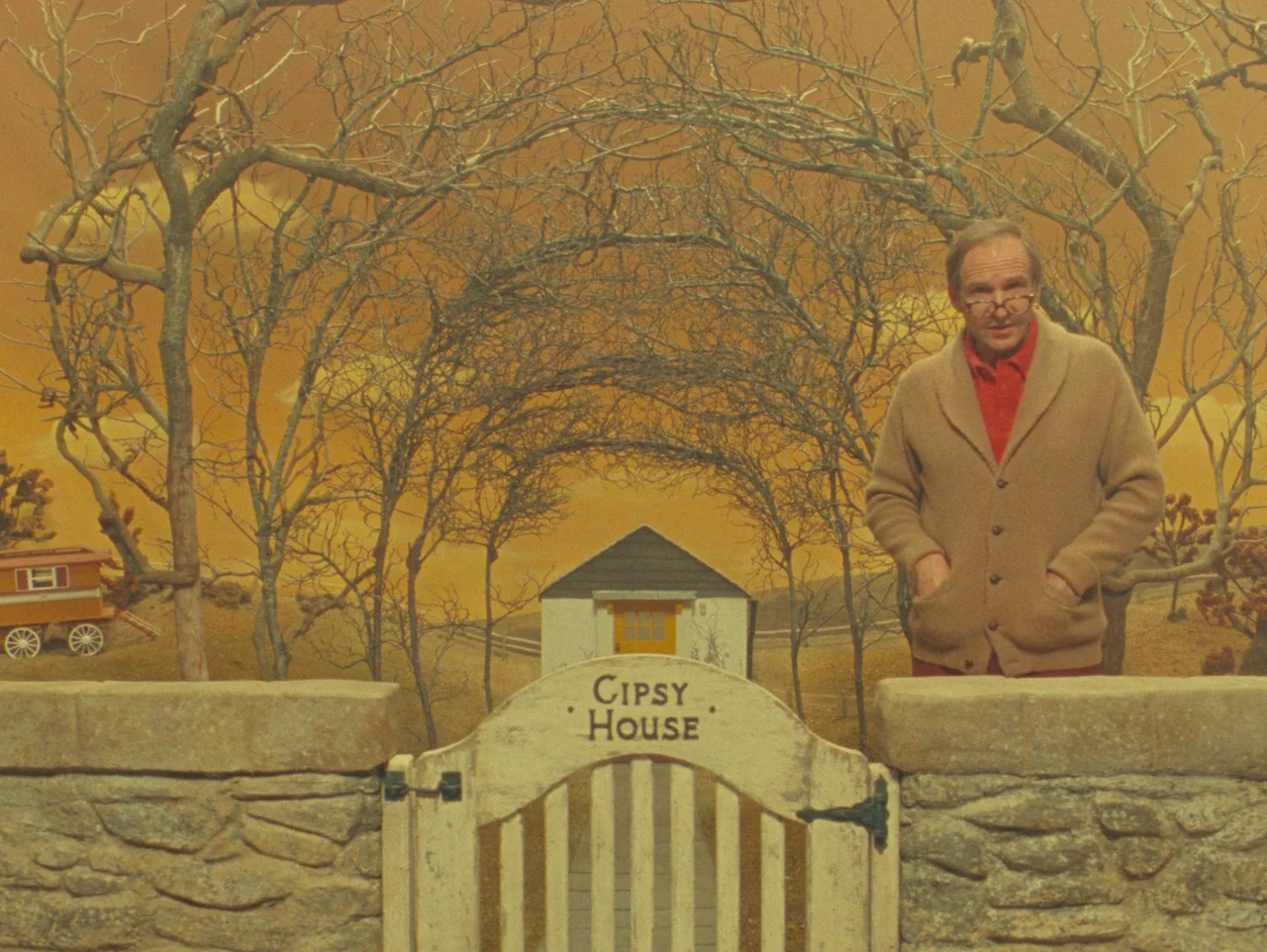 The Wonderful Story of Henry Sugar (2023) movie still: medium — A man stands behind a low stone wall featuring a gate labeled Cipsy House. The…