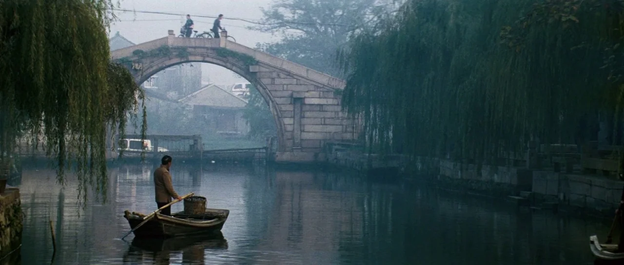 Mission: Impossible III (2006) movie still: wide — An arched stone bridge spans a narrow canal lined with weeping willow trees. A man sits in a…