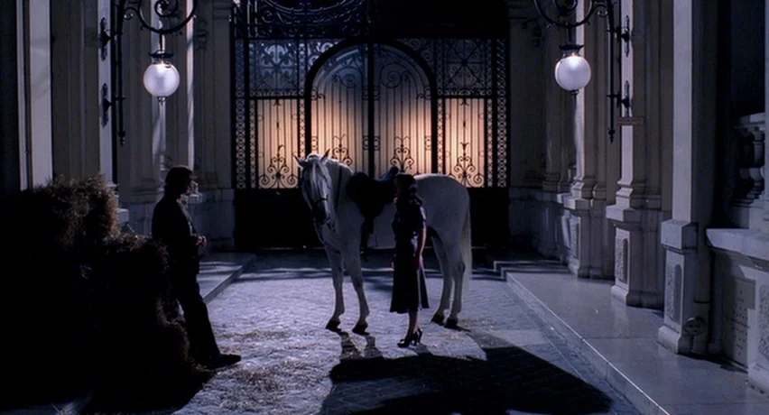 The Man Who Cried (2000) movie still: wide — An arched stone carriage entrance features ornate iron gates at the far end. A man and woman stand near…