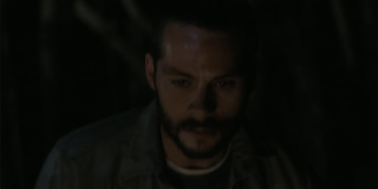 Caddo Lake (2024) movie still: closeup — A man stands in a dimly lit, nondescript interior space with a dark background. He looks down while his…