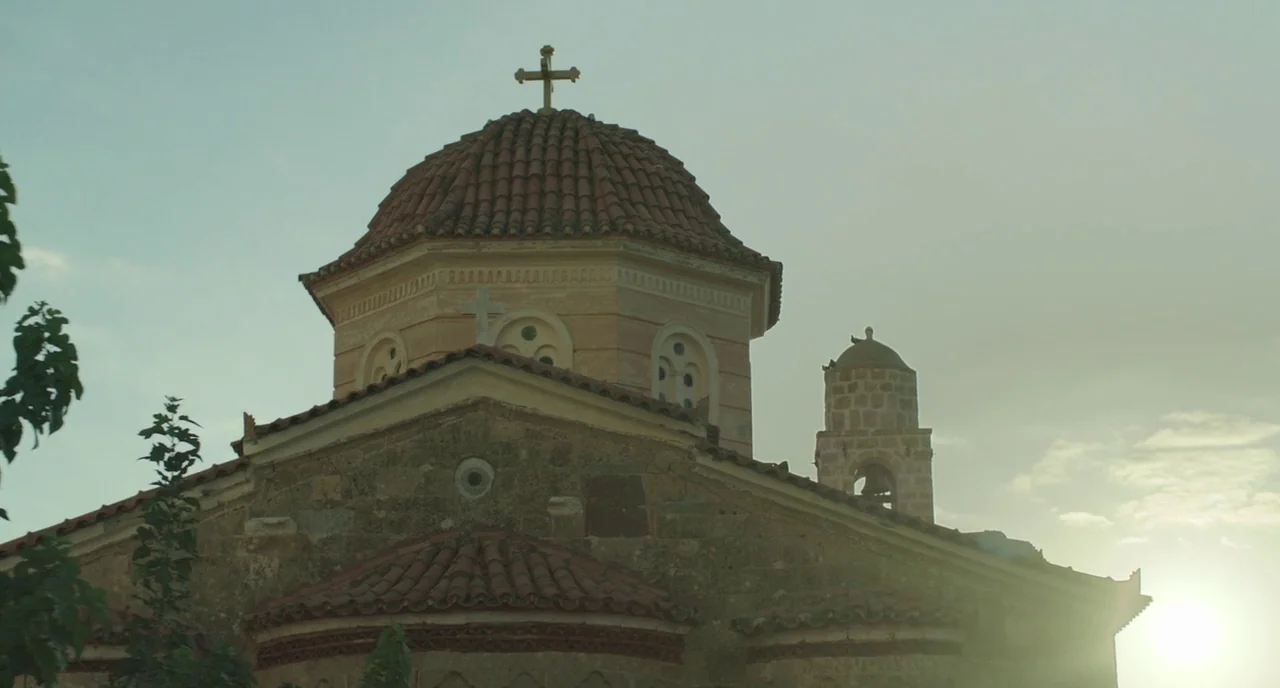 Before Midnight (2013) movie still: establishing — A stone church features a central red-tiled dome topped with a small cross. A bell tower stands…