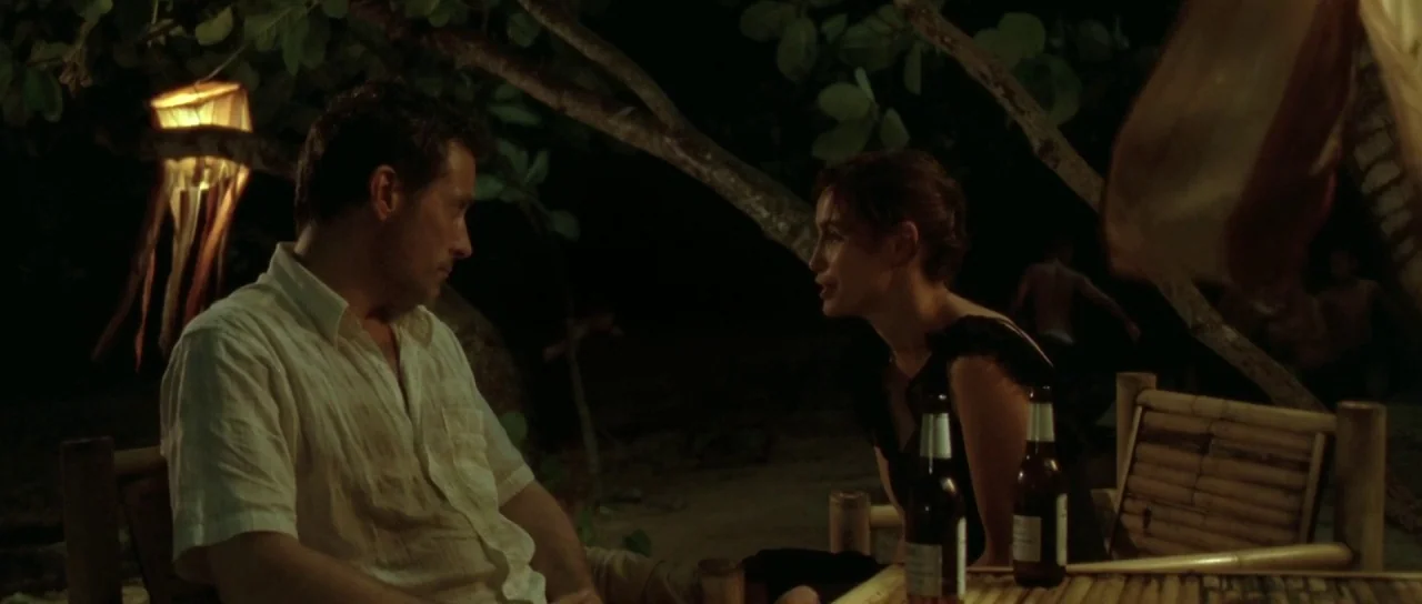 Vinyan (2008) movie still: medium — A man and woman sit at a bamboo table on a beach at night. They are talking while illuminated by warm hanging…