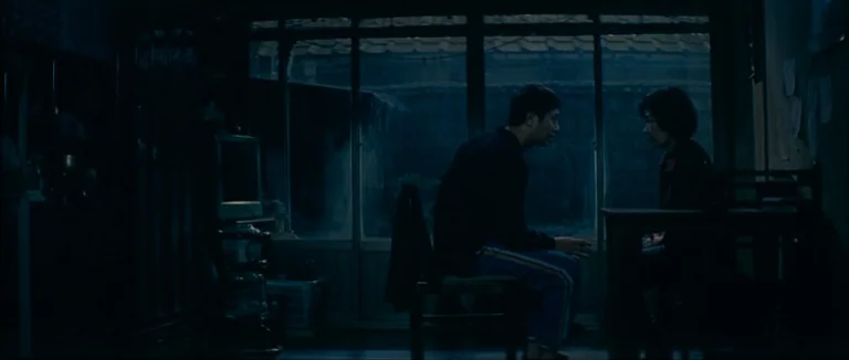 Mother (2009) movie still: wide — A dimly lit room features a sliding glass door looking out into a dark night. Two figures sit in profile at a…