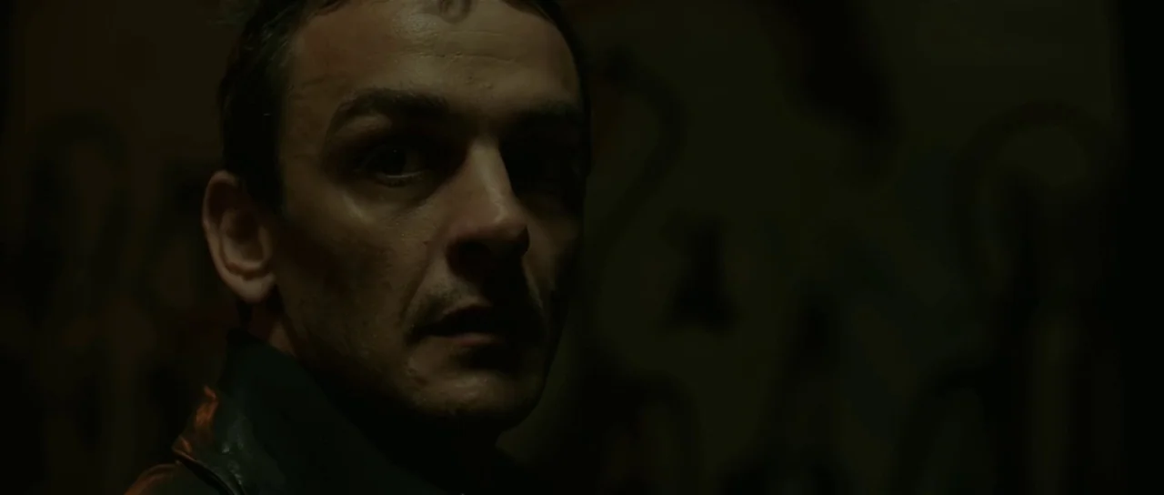 Bullhead (2011) movie still: closeup — A dimly lit interior space with dark, textured walls visible behind the subject. A middle-aged man looks…